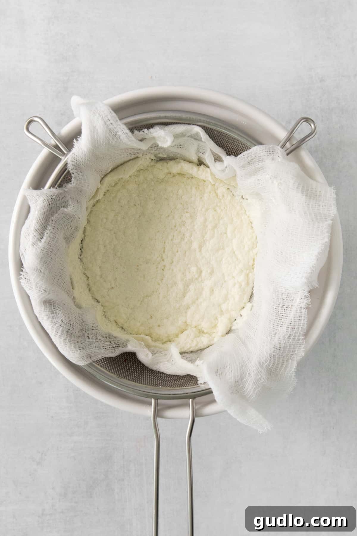 Freshly strained cheese curds, covered with a white cheesecloth, resting in a bowl over a sink, highlighting the crucial step of draining excess whey.