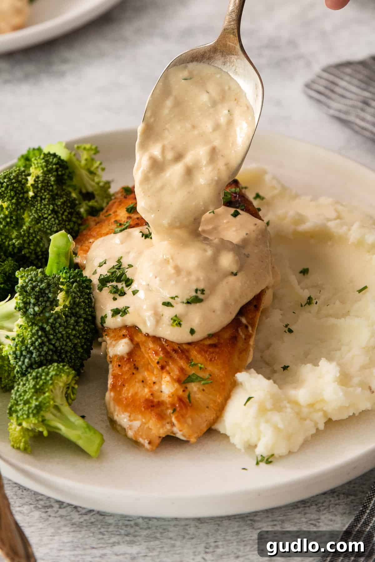 Crispy Garlic Chicken Delight 2 A person pouring a rich, creamy sauce over golden-brown chicken and vibrant green broccoli florets on a pristine white plate, promising a delightful meal.