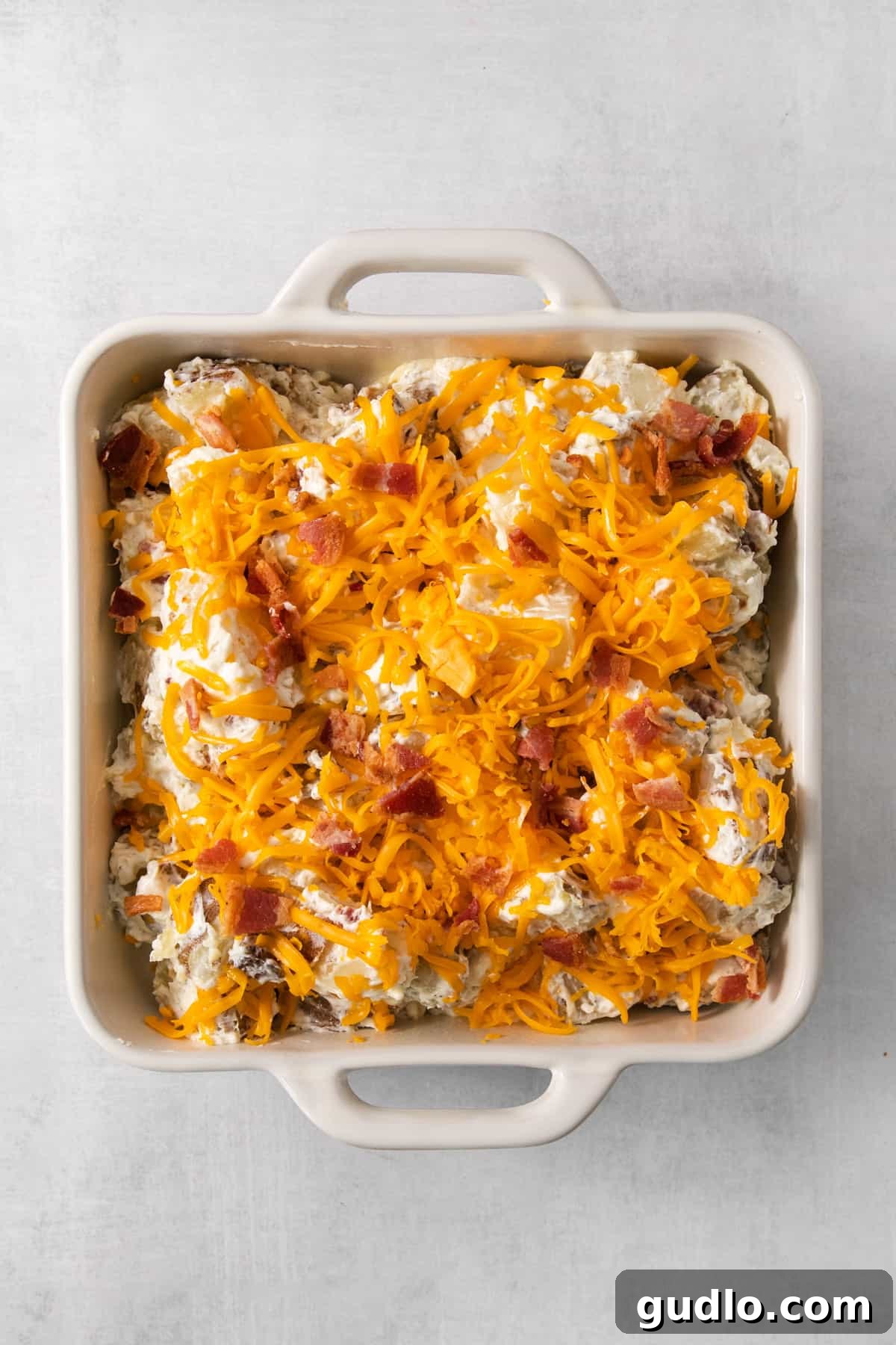 Ultimate Loaded Potato Bake 5 A casserole dish filled with a creamy potato mixture, topped generously with crispy bacon pieces and shredded cheddar cheese, ready for baking.