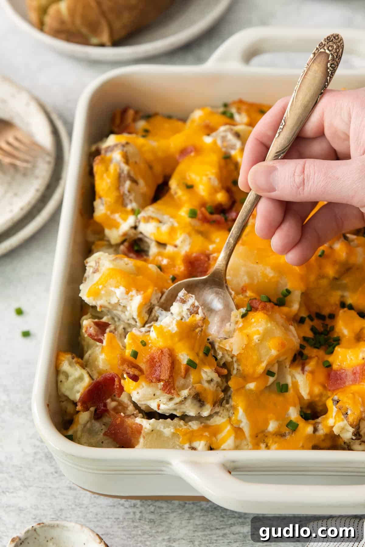 Ultimate Loaded Potato Bake 6 A person scooping a generous portion of hot, cheesy Loaded Baked Potato Casserole from a white baking dish with a large spoon.