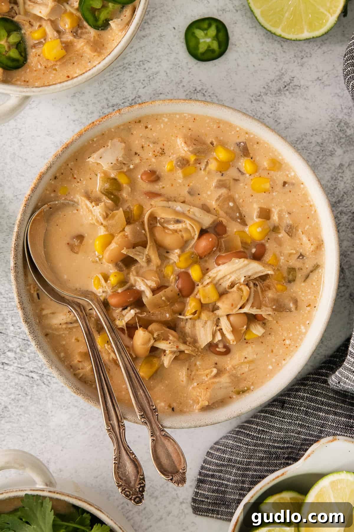 Creamy Slow Cooker White Chicken Chili 4 A bowl of vibrant Mexican chicken chowder, garnished with fresh lime wedges and sliced jalapeños, resting on a white surface.