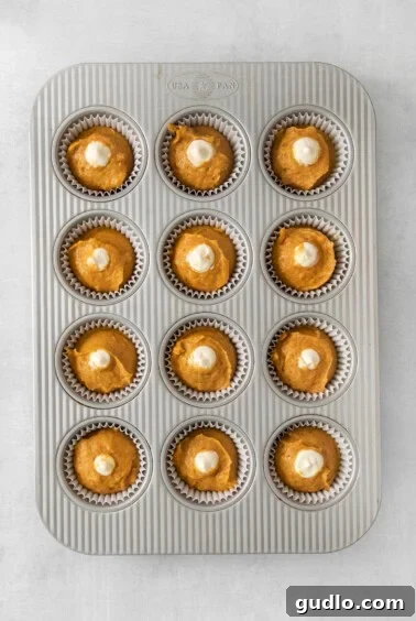 Cream Cheese Swirl Pumpkin Muffins 13 Close-up of unbaked pumpkin cupcakes nestled in a muffin tin, each topped with a dollop of cream cheese filling, ready for the oven.