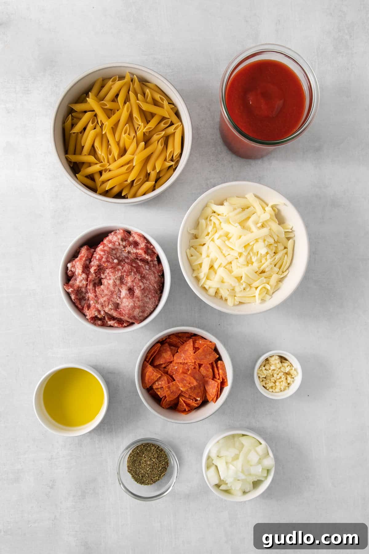 A collection of fresh ingredients for pizza casserole, including pasta, ground meat, and tomatoes, arranged on a white surface.