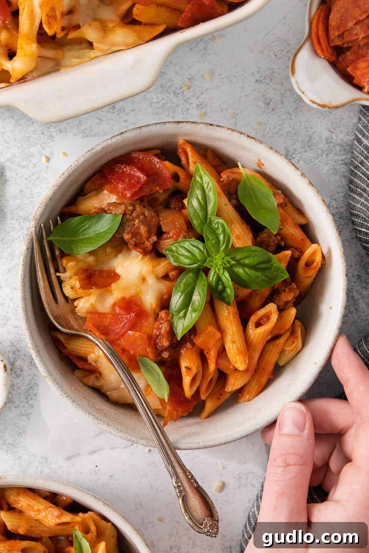 A person holding a bowl filled with pepperoni pizza casserole, garnished with fresh basil, ready to eat.