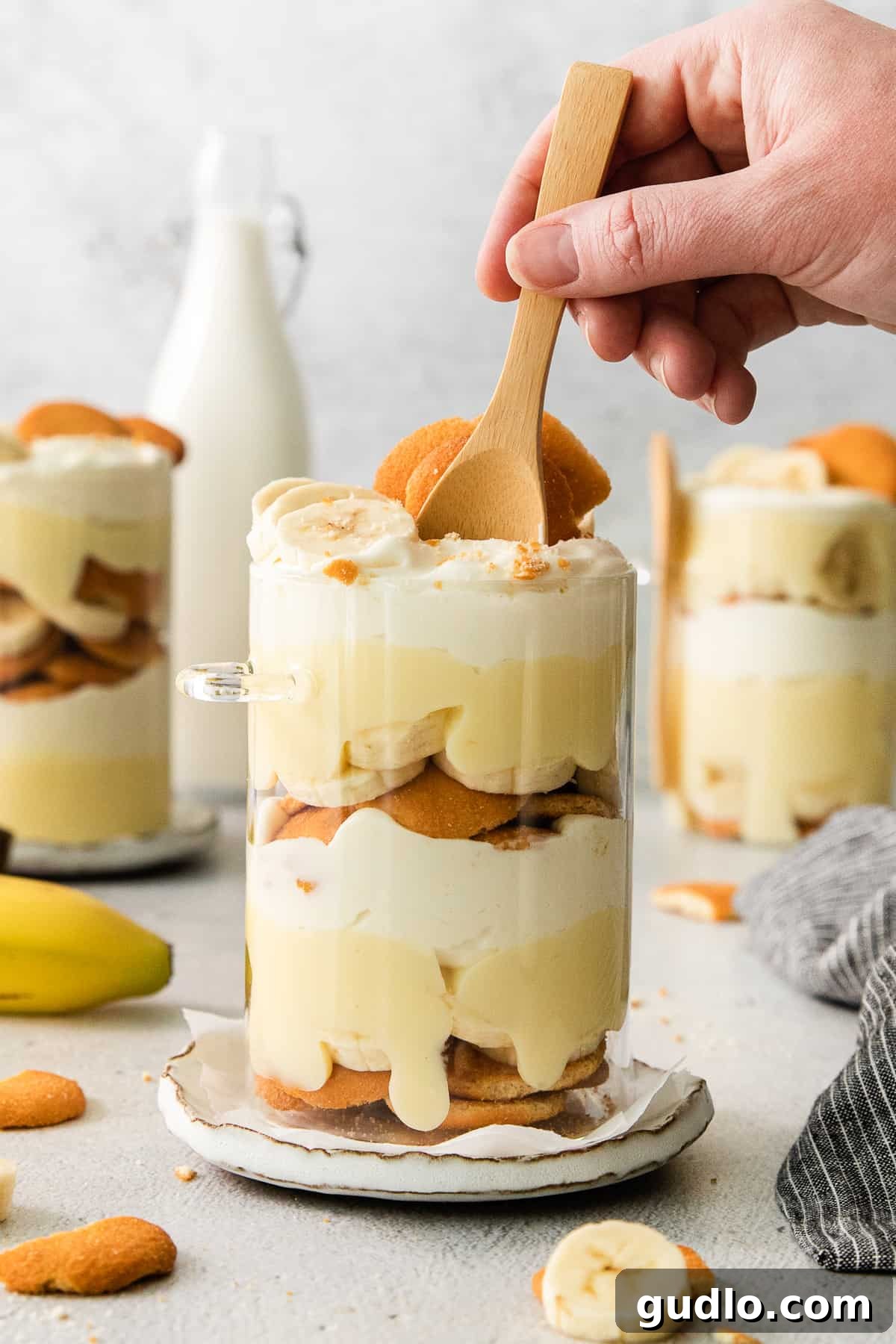 Decadent Banana Cream Cheese Pudding 6 A person holding a wooden spoon poised over a beautifully layered dessert in a glass dish, ready to serve.