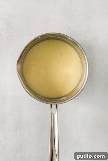 Decadent Banana Cream Cheese Pudding 10 A close-up of a metal spoon with liquid being poured into a mixture, demonstrating the tempering process for egg yolks.