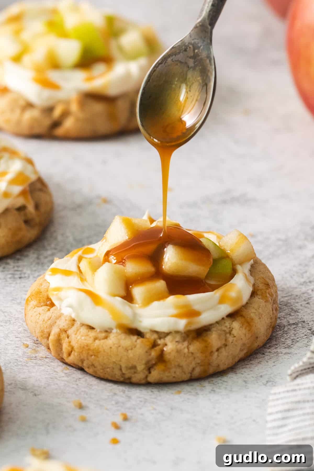 Caramel Apple Cheesecake Cookies 3 A spoon delicately drizzling caramel sauce over a freshly frosted cream cheese caramel apple cookie.