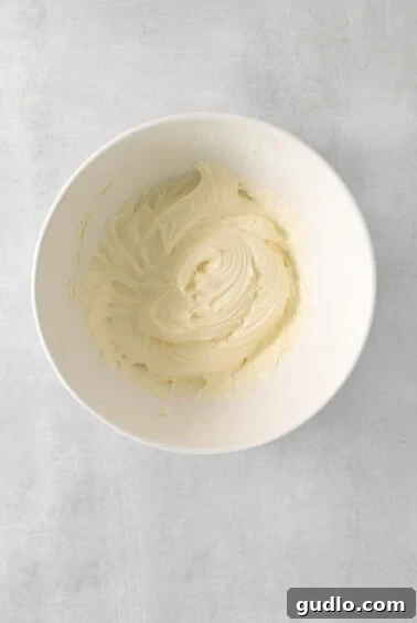 A white bowl containing smooth, creamy cream cheese frosting, ready for application, with a hand mixer nearby.
