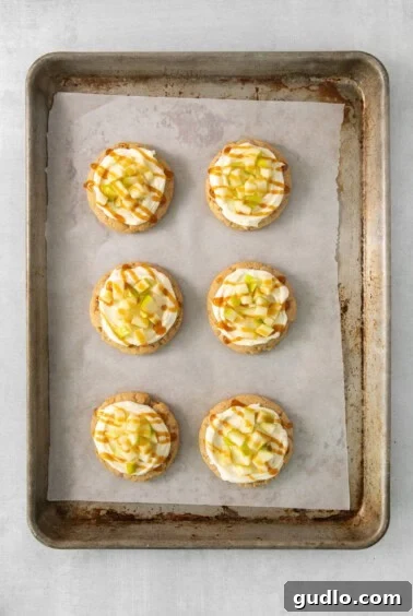 A baking tray showcasing several finished cream cheese caramel apple cookies, decorated with frosting and apple pieces.