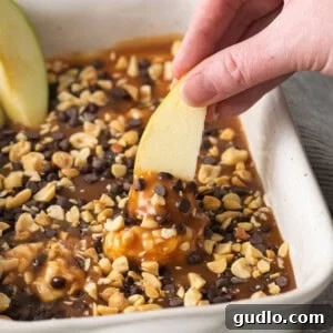 Caramel Apple Cheesecake Cookies 4 A person dipping an apple slice into a creamy, golden caramel apple dip in a baking dish.