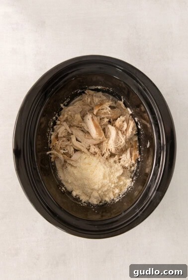 A bowl of shredded chicken in a slow cooker, mixed with the creamy sauce and mushrooms, ready for the final step with Parmesan cheese.