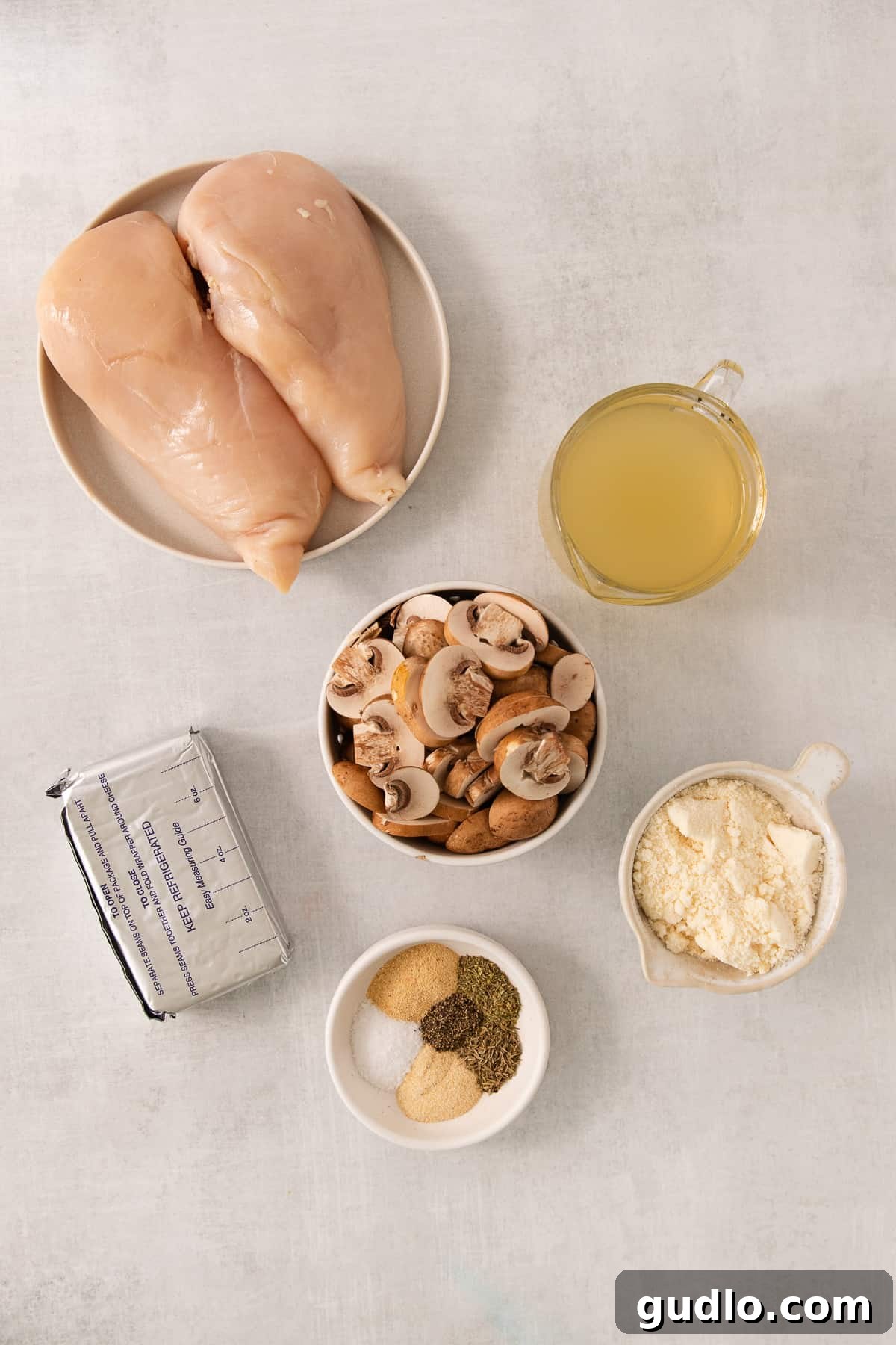 All the fresh ingredients for making slow cooker cream cheese chicken, including whole chicken breasts, a block of cream cheese, grated Parmesan, sliced crimini mushrooms, and a selection of dried herbs and spices, neatly arranged on a white kitchen counter.