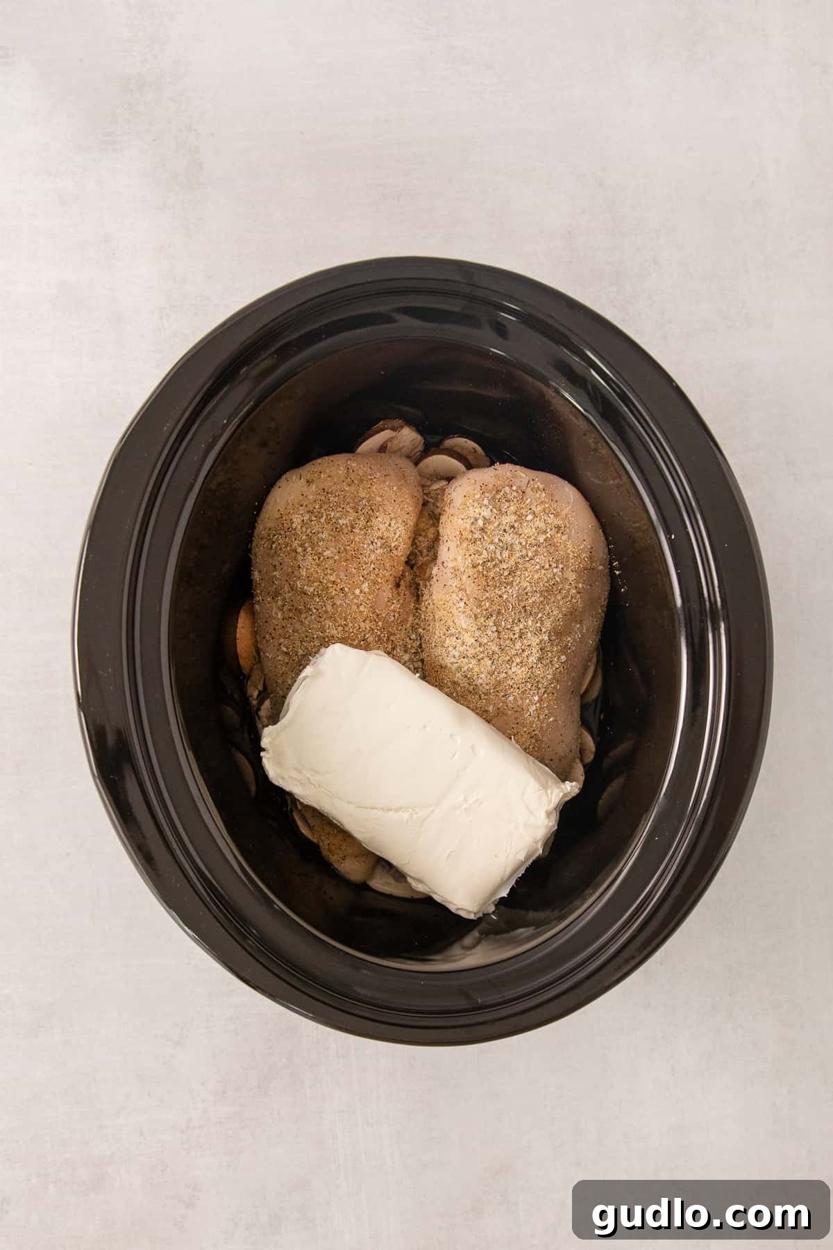 A black crock pot filled with raw boneless chicken breasts, a block of cream cheese, and sliced mushrooms, ready for the slow cooking process to begin.