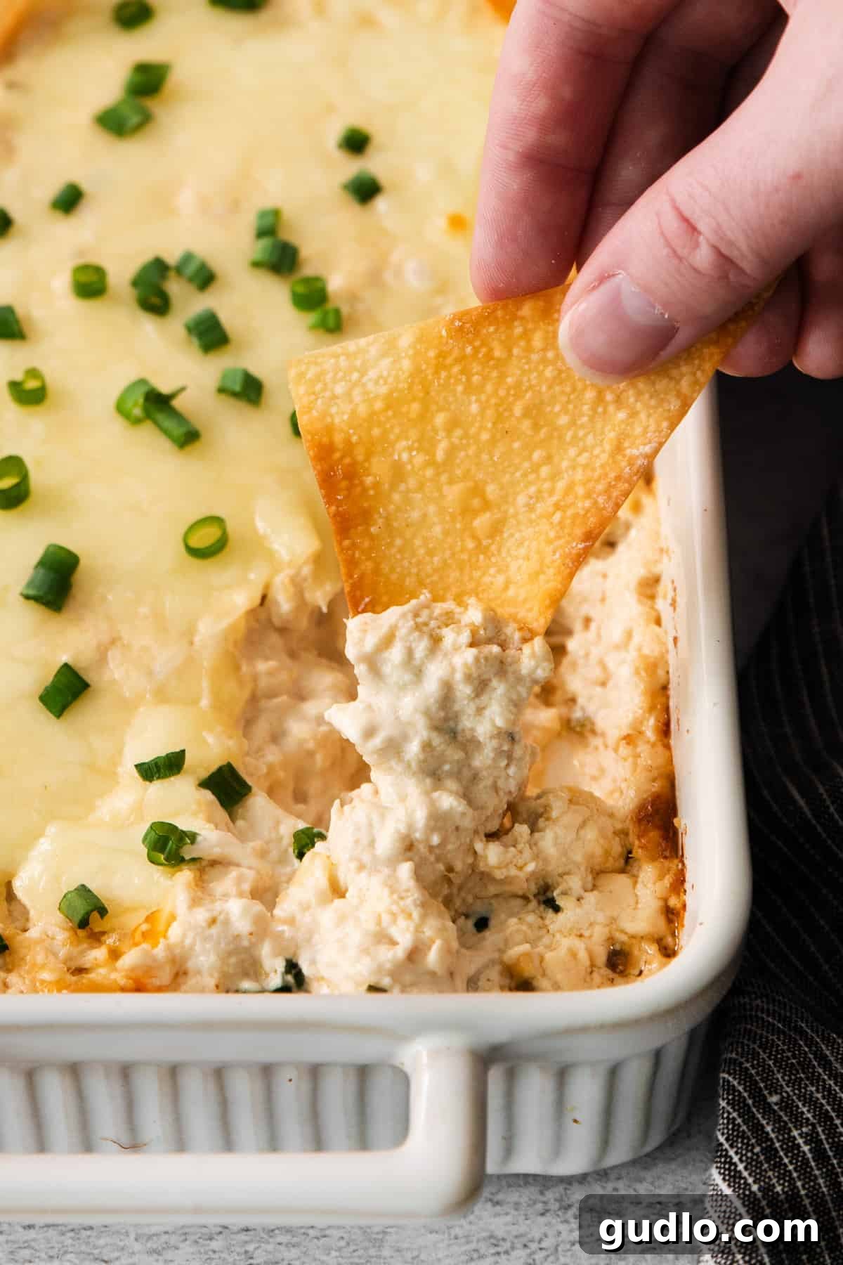 Deconstructed Crab Rangoon Dip 2 A hand dipping a crispy wonton chip into a creamy, golden-brown baked crab rangoon dip, served in a white baking dish.