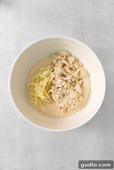 A white bowl with a creamy mixture, now containing flaked crab meat and shredded mozzarella cheese, ready to be stirred.