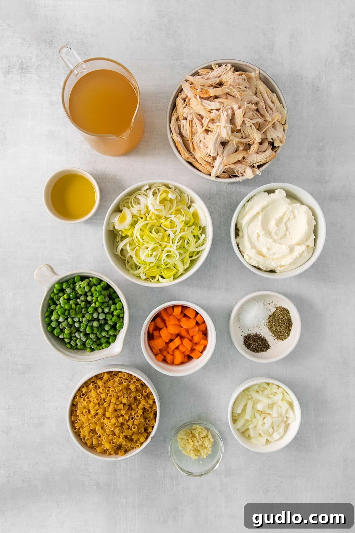 Golden Cream Chicken 3 A collection of fresh ingredients laid out on a rustic wooden surface, including onions, carrots, leeks, garlic, chicken broth, ditalini pasta, shredded chicken, and a container of ricotta cheese, all ready for making creamy chicken soup.