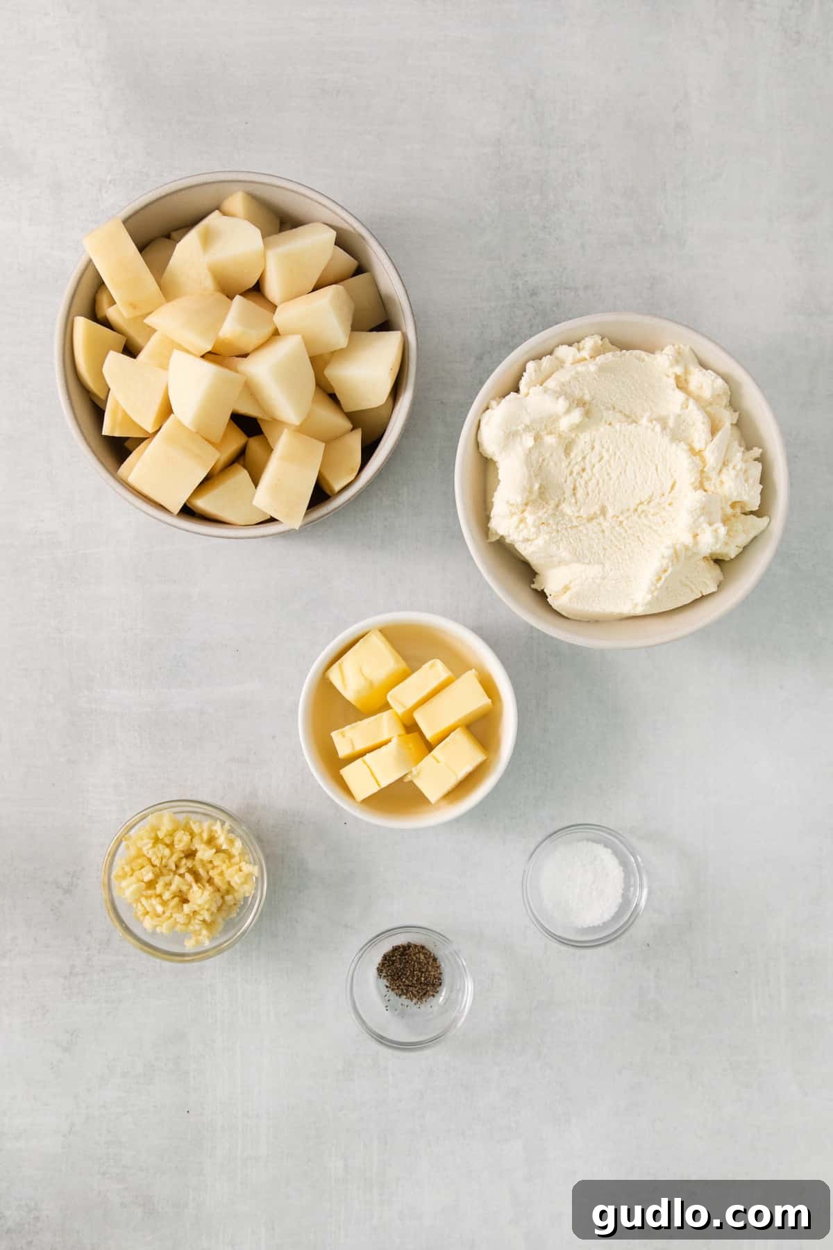 Luxurious Garlic Mashed Potatoes 3 A collection of fresh ingredients: ricotta cheese, butter sticks, whole garlic cloves, and salt flakes, arranged on a gray background.