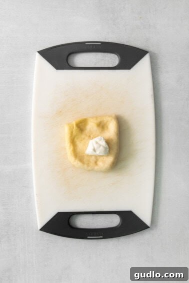 A piece of flattened dough on a wooden cutting board with a chunk of cream cheese placed in the center.