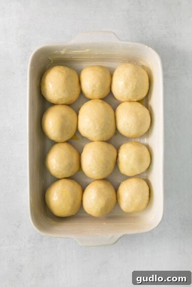 A white baking dish filled with neatly arranged, unbaked dough balls, brushed with egg wash and ready for the second rise.