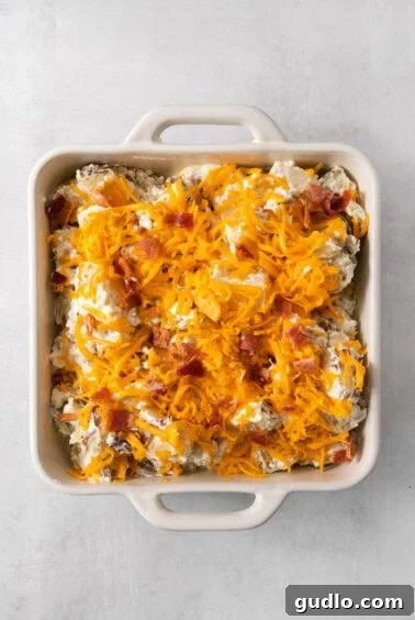 Decadent Twice-Baked Potato Bake 11 A casserole dish filled with a creamy potato mixture, generously topped with shredded cheese and crispy bacon, ready for baking.