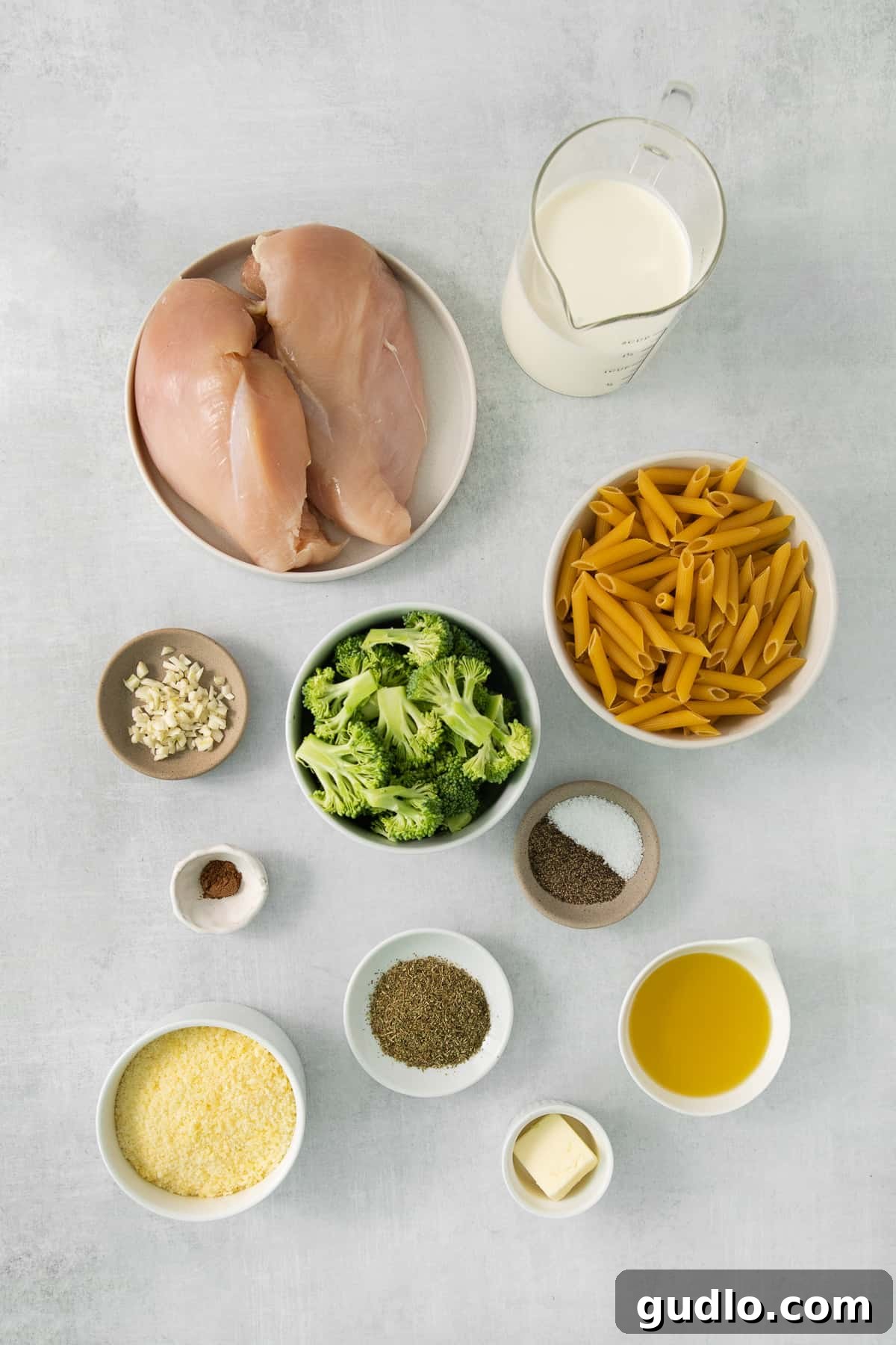 Arrangement of fresh chicken, broccoli, heavy cream, garlic, and penne pasta on a grey surface, ready for cooking.