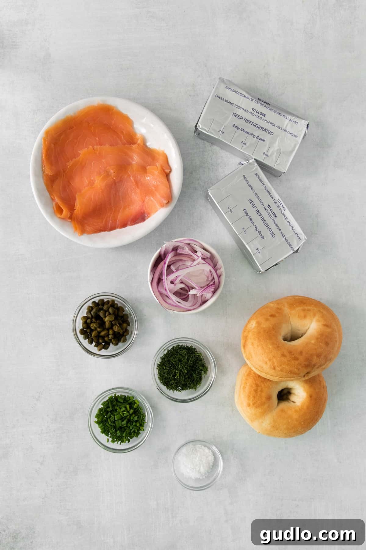 Essential ingredients for a smoked salmon bagel sandwich laid out on a clean surface: smoked salmon, red onion, capers, cream cheese, and fresh dill.