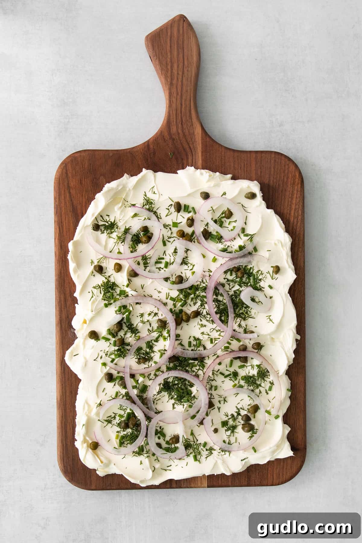 A close-up shot of the cream cheese board, showing a generous spread of cream cheese topped with fresh dill, chives, red onions, and capers, before the salmon is added.
