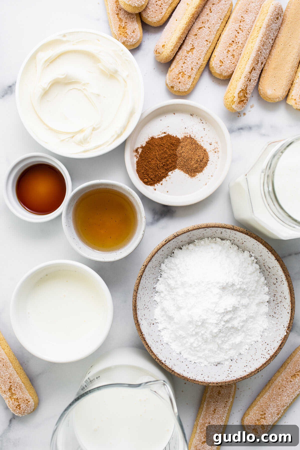 A collection of fresh ingredients for Eggnog Tiramisu, including eggnog, rum, ladyfingers, and spices, arranged on a marble surface.