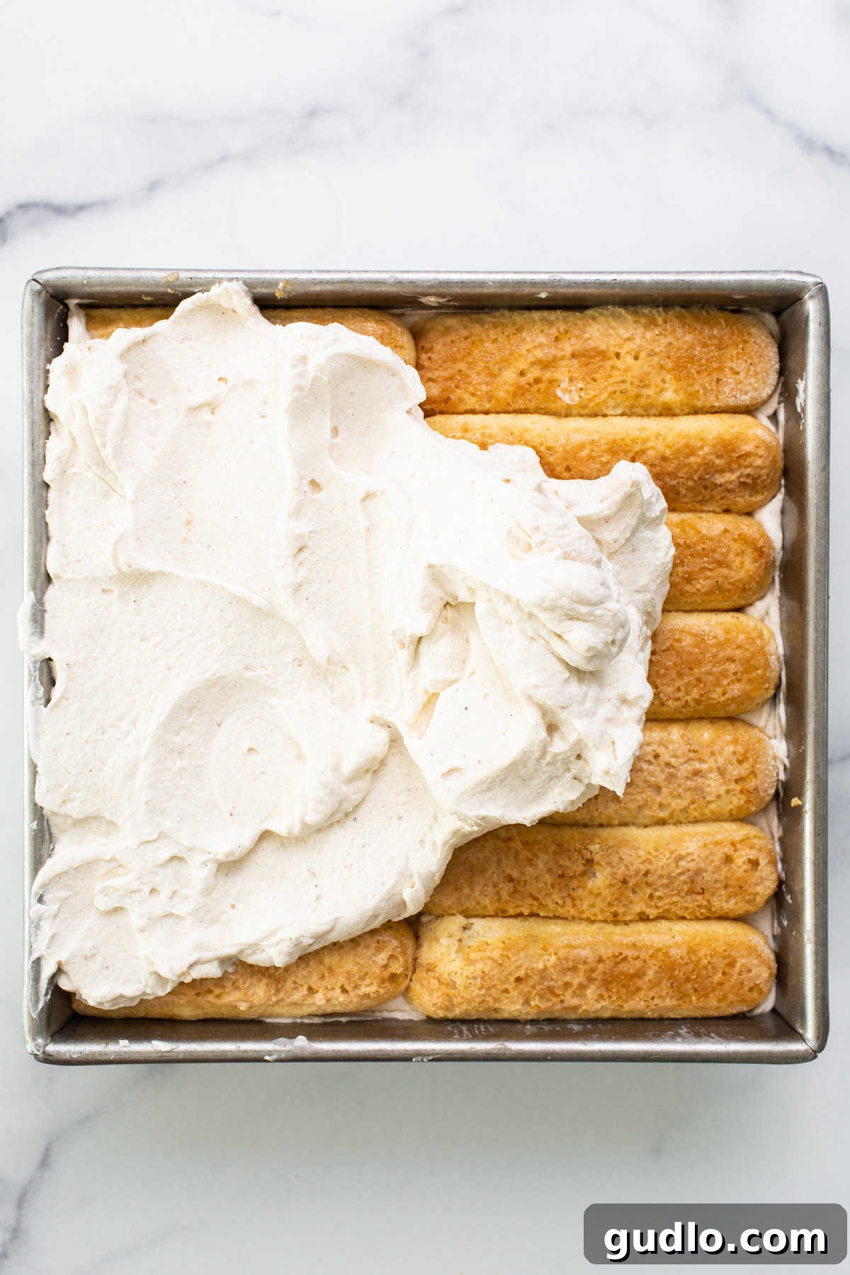 A baking pan filled with the assembled layers of Eggnog Tiramisu, topped with a generous layer of creamy mascarpone, ready for chilling.