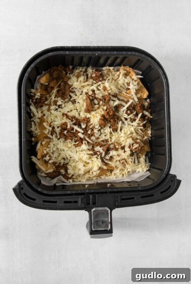 No-Fuss Air Fryer Nachos 10 An air fryer basket generously filled with tortilla chips, seasoned taco meat, and shredded cheese, ready for cooking.