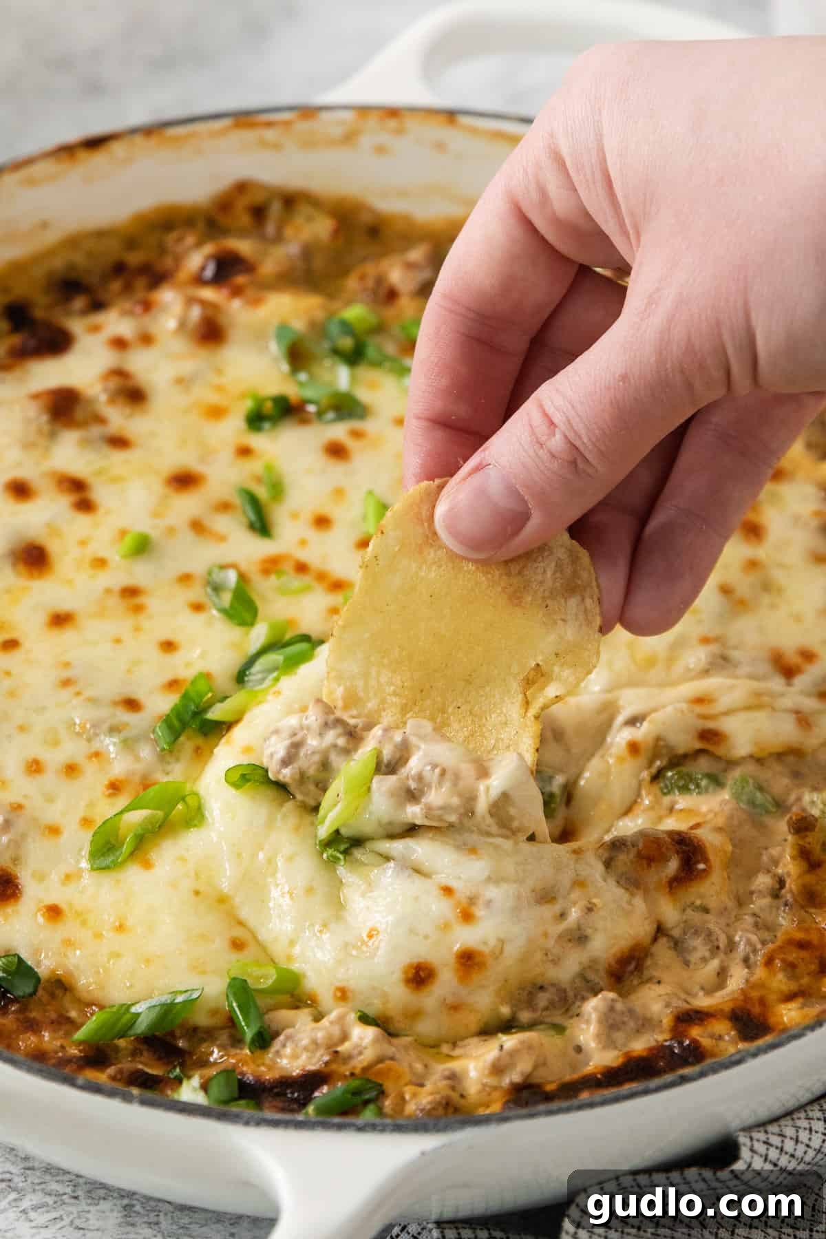 Decadent Philly Cheesesteak Dip 5 A hand dipping a crispy chip into the warm, gooey Philly Cheesesteak Dip.