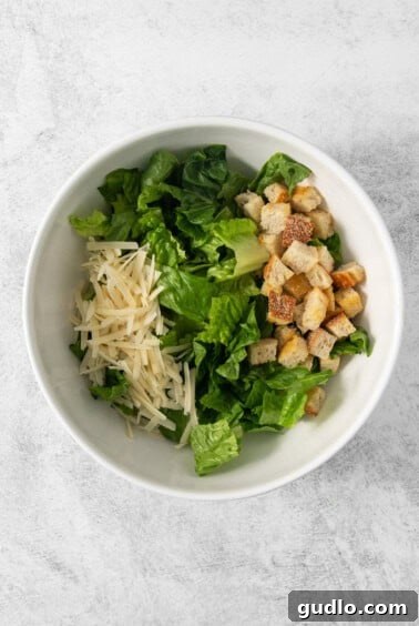 A large white bowl containing crisp romaine lettuce, crunchy croutons, and generous amounts of Parmesan cheese, waiting for the dressing.
