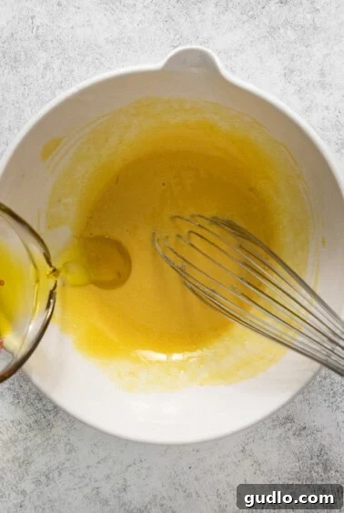 A whisk actively blending a yellow liquid in a white bowl, demonstrating the thickening of the dressing as olive oil is gradually added.