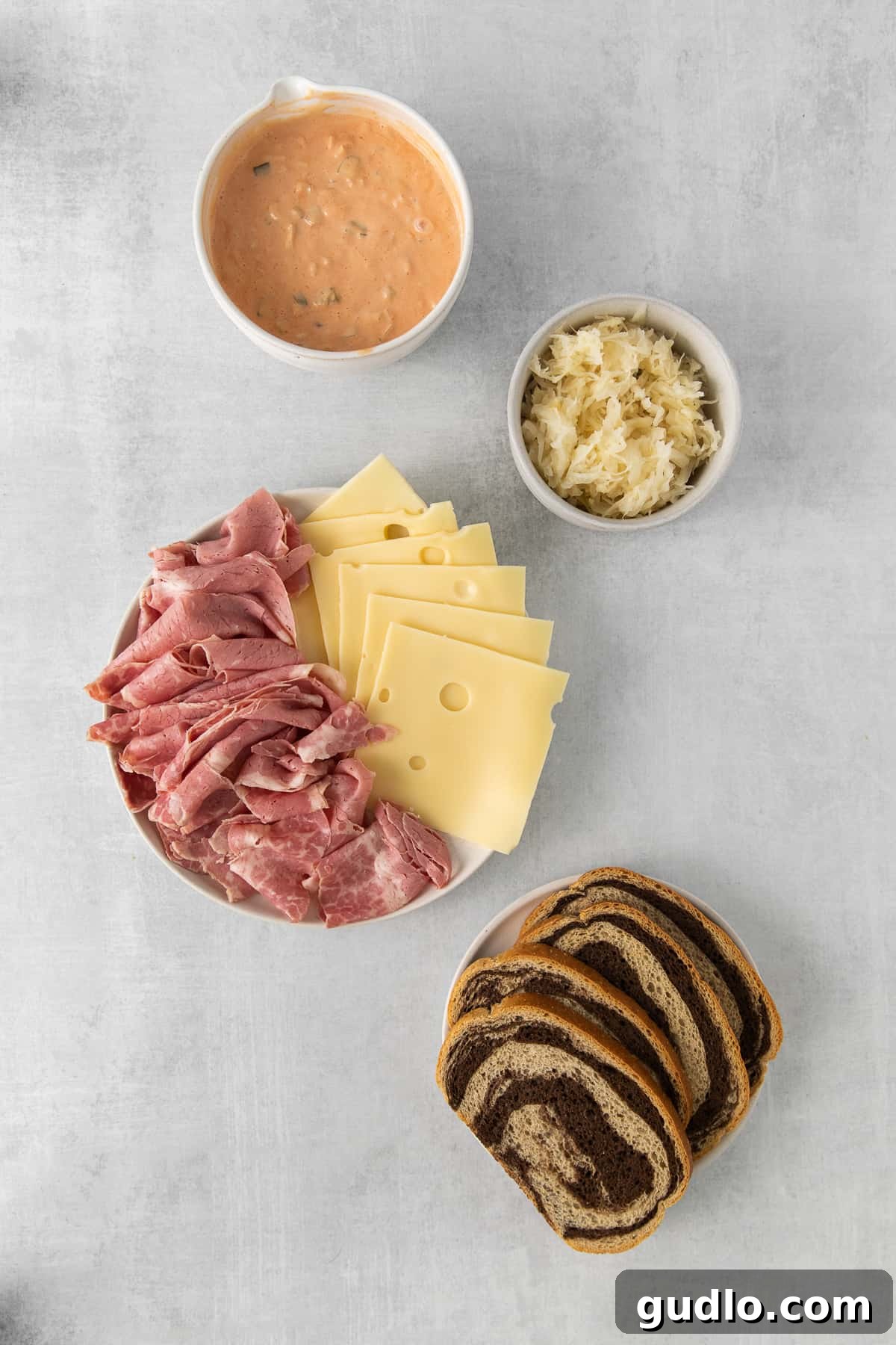 An array of ingredients for a Reuben sandwich laid out: sliced ham, Swiss cheese, marbled rye bread, and a bowl of Thousand Island dressing.