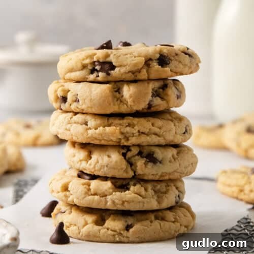 A stack of five egg-free chocolate chip cookies on a white surface, with more cookies and blurred bottles in the background.