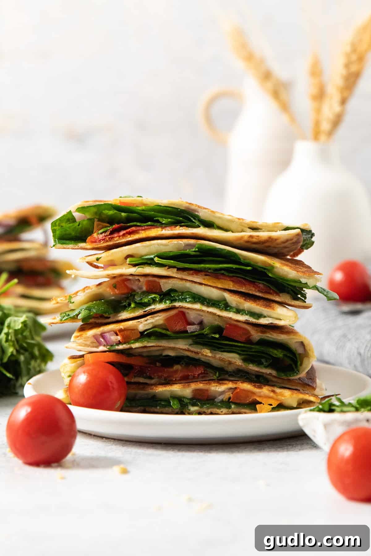 Mediterranean Sunshine Quesadilla 2 A stack of sliced vegetable quesadillas on a white plate, surrounded by cherry tomatoes and fresh spinach, with a blurred background of vases and wheat stalks.