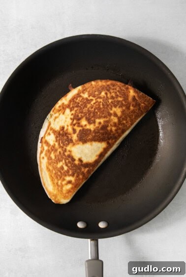 A browned, folded quesadilla is cooking in a black non-stick frying pan.