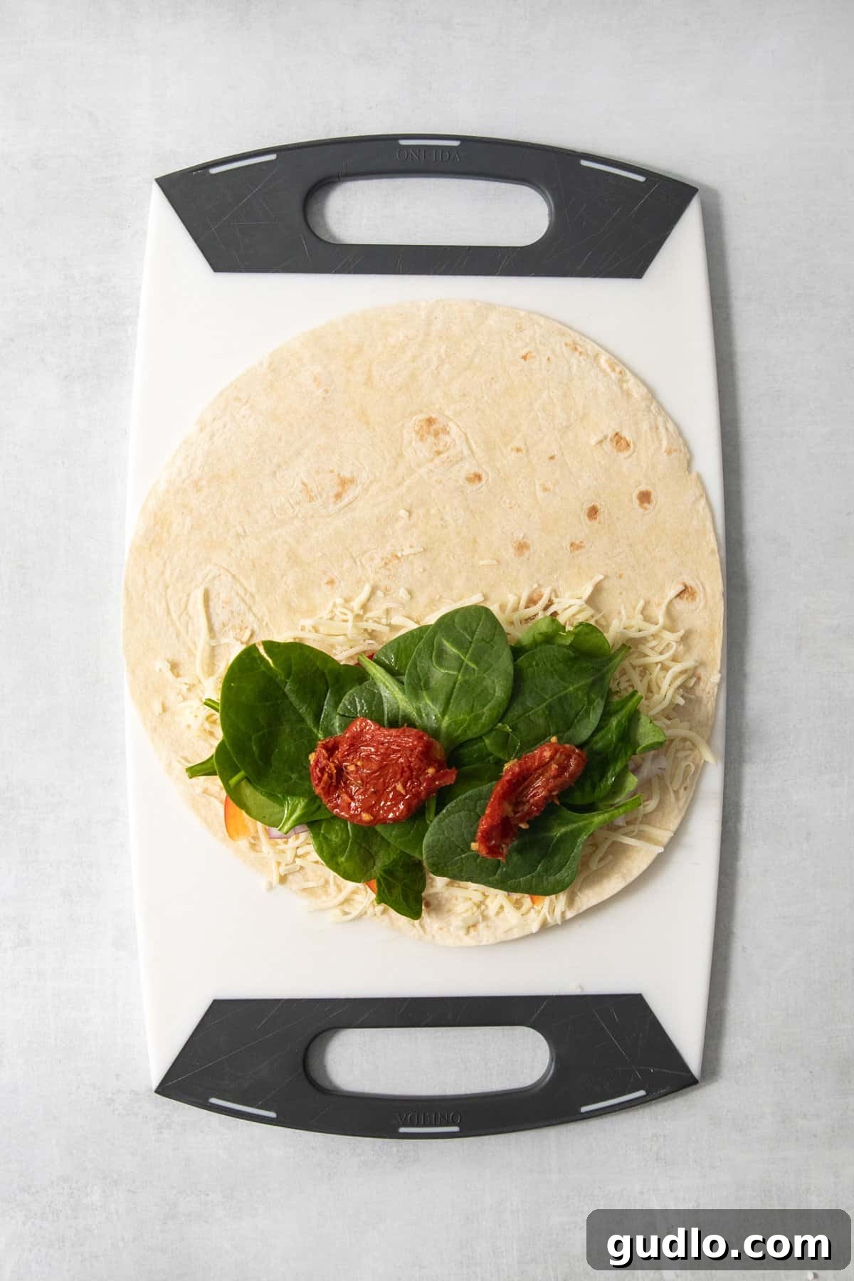 Mediterranean Sunshine Quesadilla 3 A tortilla with shredded cheese, leafy greens, and sun-dried tomatoes placed on a cutting board.