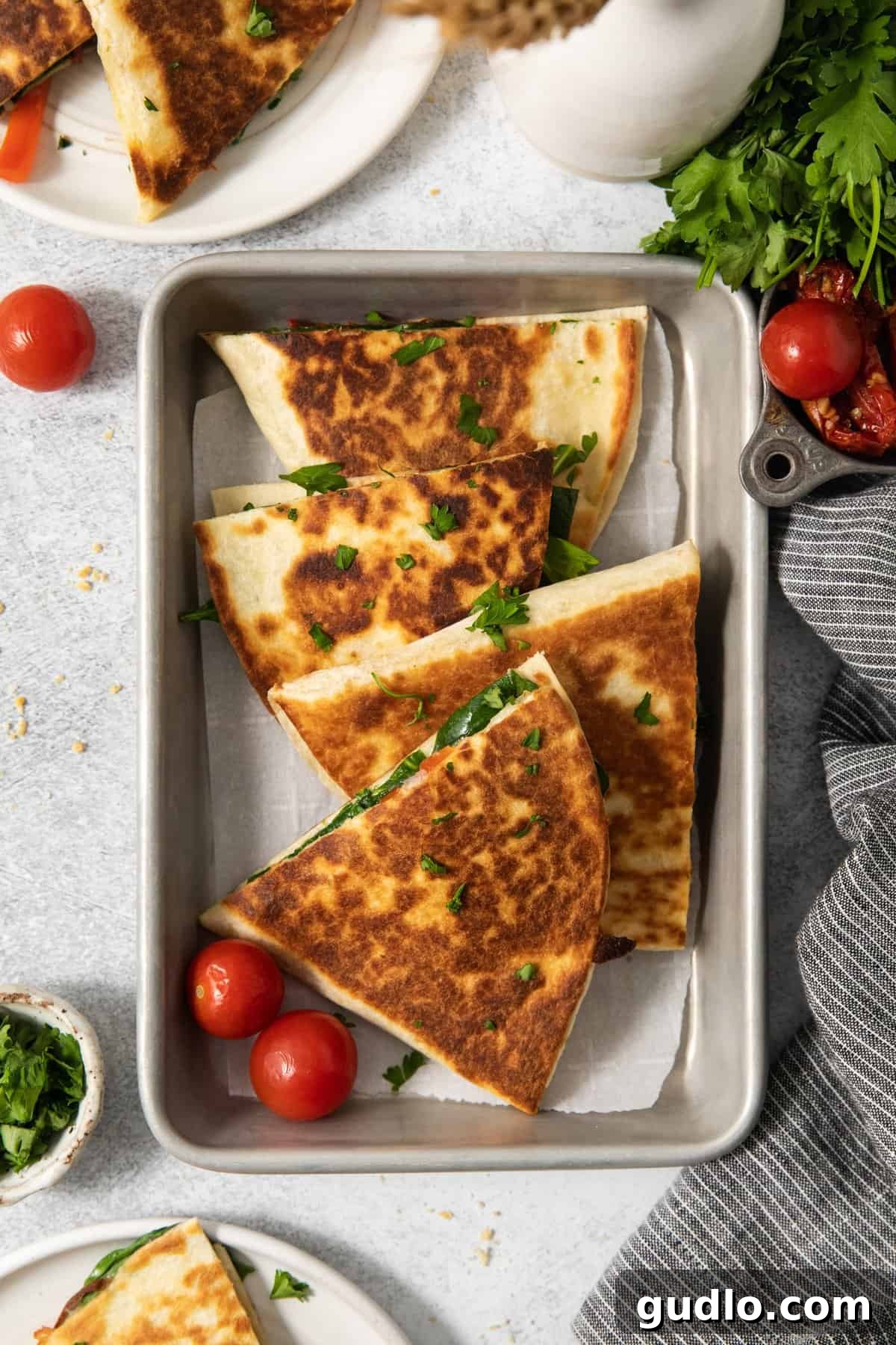 Mediterranean Sunshine Quesadilla 4 A metal tray with browned quesadilla slices garnished with chopped herbs, accompanied by whole cherry tomatoes. Fresh greens and tomatoes are visible nearby.
