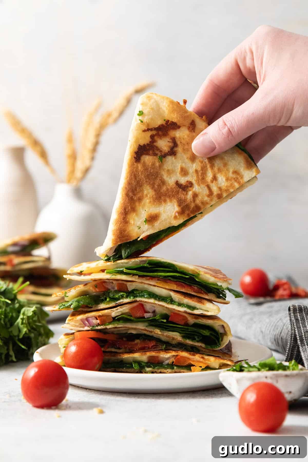 Mediterranean Sunshine Quesadilla 6 A hand lifting a slice from a stack of quesadillas filled with vegetables, with tomatoes, greens, and decorative items in the background.
