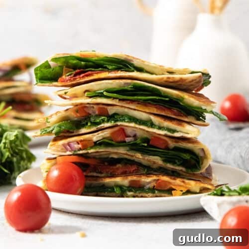 A plate of stacked quesadilla wedges filled with spinach, tomatoes, cheese, and onions. Two cherry tomatoes sit beside the plate.