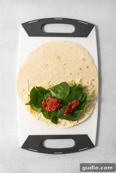 A tortilla with shredded cheese, leafy greens, and sun-dried tomatoes placed on a cutting board.