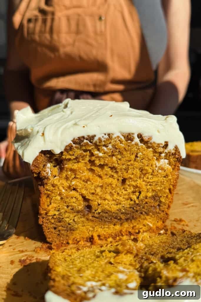 A close-up of a sliced loaf cake with white frosting, sitting on a wooden cutting board; a person in a tan apron stands in the background.