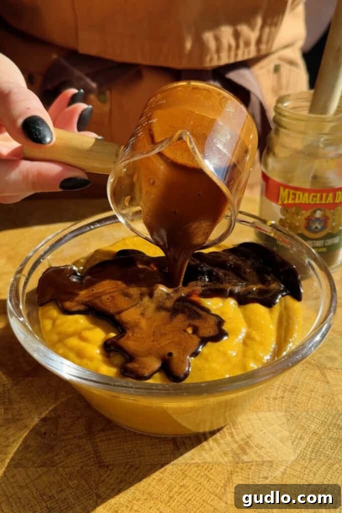 A hand pours thick chocolate sauce from a glass cup onto a bowl of yellow pudding. A jar labeled "Medaglia D’Oro" is visible in the background.