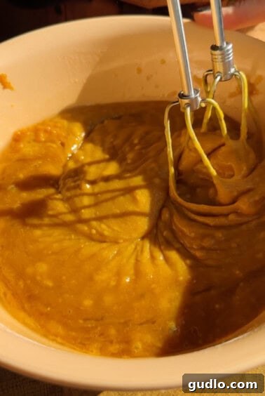 A close-up of a mixing bowl containing thick orange batter being mixed with an electric hand mixer.