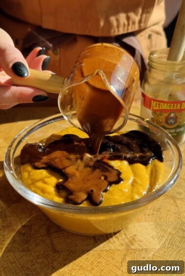 A hand pours thick chocolate sauce from a glass cup onto a bowl of yellow pudding. A jar labeled "Medaglia D’Oro" is visible in the background.