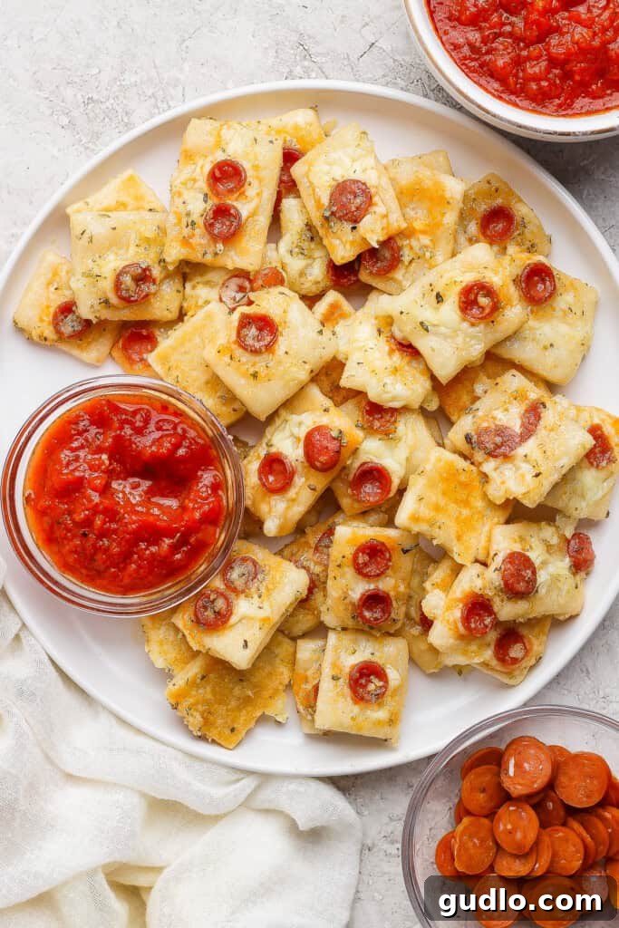 Protein Packed Pepperoni Pizza Bites 2 A plate of rectangular cheesy bread bites topped with mini pepperoni slices, served with bowls of marinara sauce and extra mini pepperoni on the side.