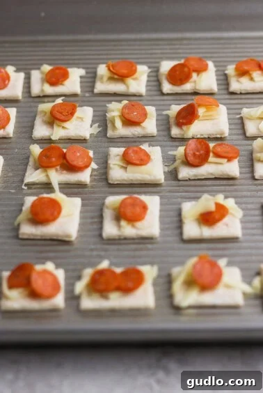 Protein Packed Pepperoni Pizza Bites 8 A baking sheet with rows of square crackers topped with shredded cheese and small pepperoni slices, ready to be baked.