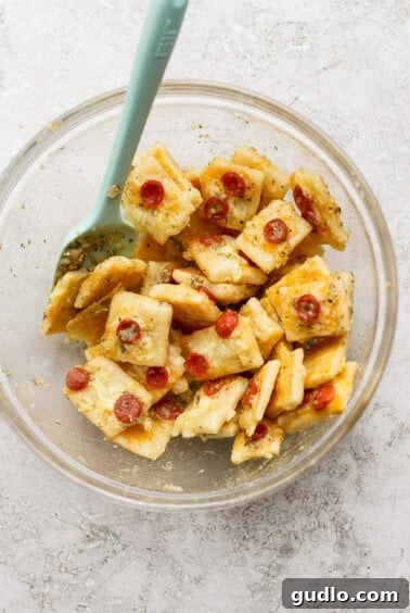 Protein Packed Pepperoni Pizza Bites 10 A glass bowl filled with seasoned crackers, shredded cheese, and mini pepperoni slices, mixed with a light blue spatula.