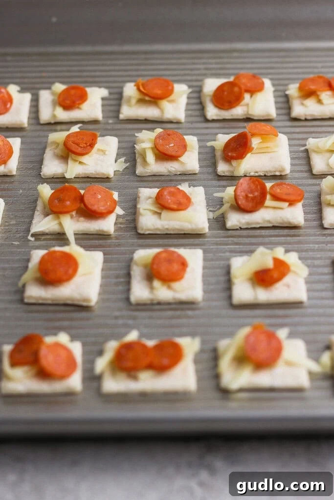 Protein Packed Pepperoni Pizza Bites 4 A baking sheet with rows of square crackers topped with shredded cheese and small pepperoni slices, ready to be baked.